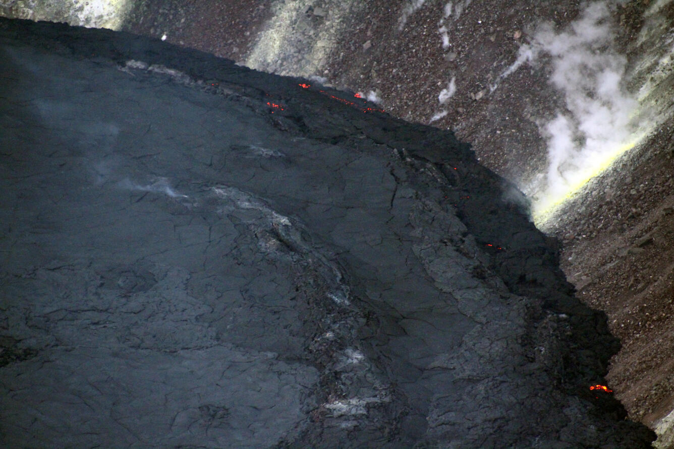 Telephoto view of dark stagnant lava lake margin with active lava oozing between the edge and the rocky crater wall