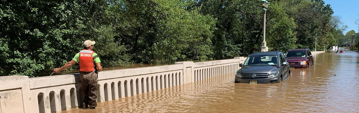 Image shows a USGS scientist in a PFD walking along a flooded roadway fence