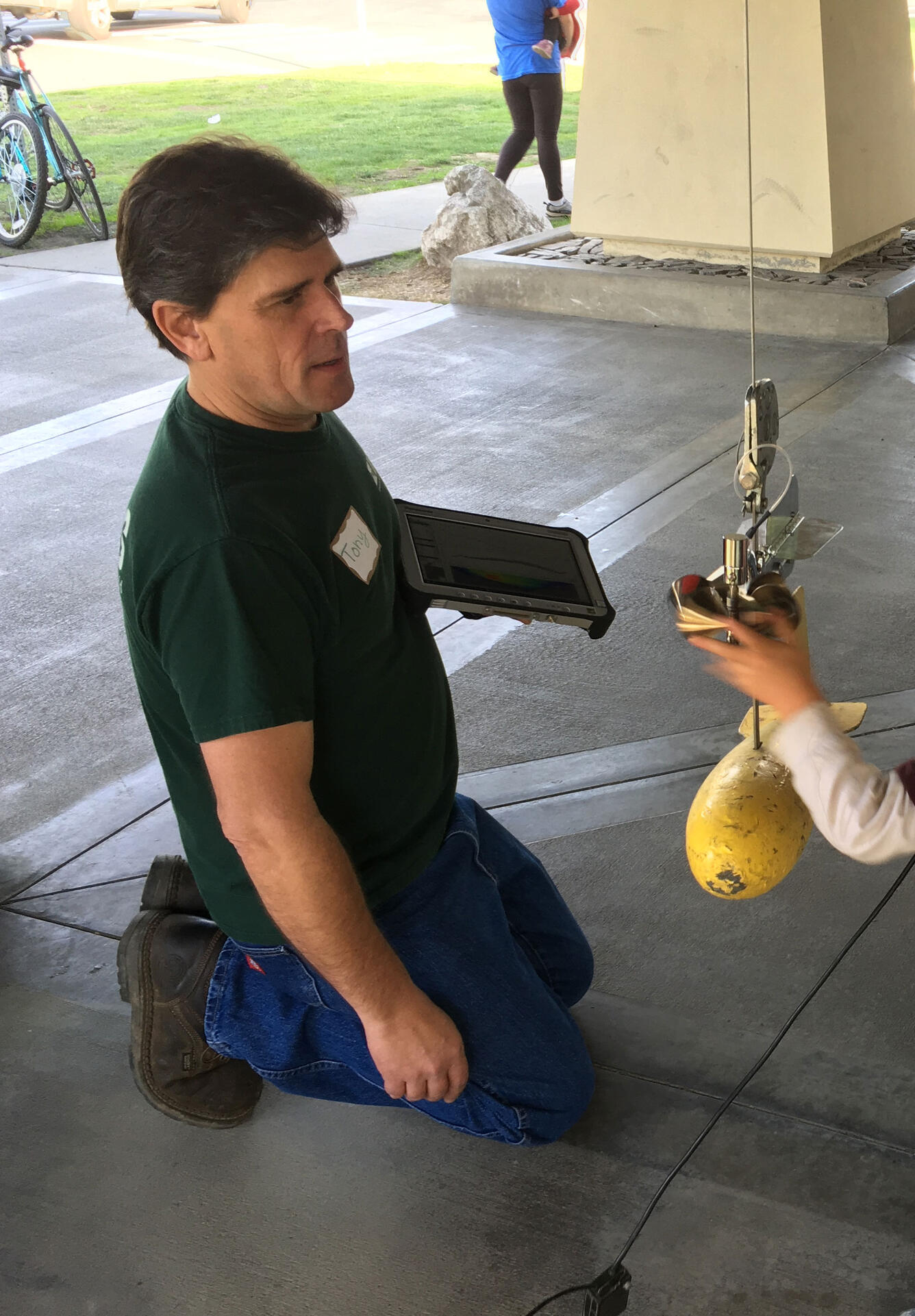 A USGS employee kneels near a small, yellow, torpedo-shaped device that is hanging in front of him.