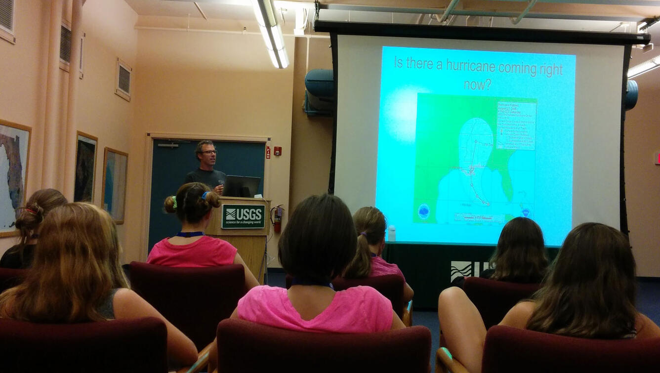 A man stands at a podium near a screen with an image projected onto it, he is talking to a group of kids.