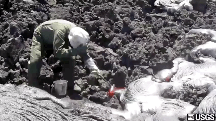A person scoops lava from a flow with a rock hammer while wearing protective gear and shielding their face.