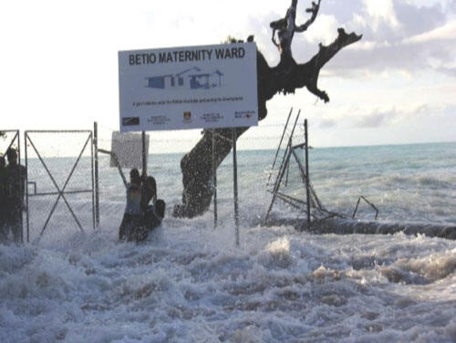 Waves crash over a seawall and mangled fencing with people hanging onto the fence.