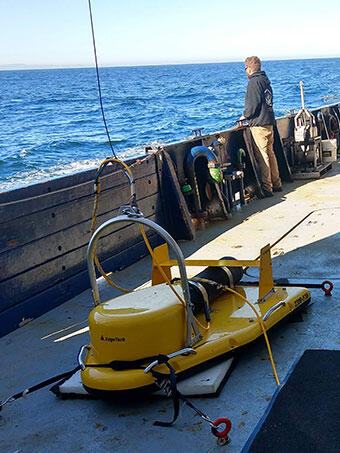 An instrument sits on the deck of a ship, attached to a cable, with a man standing behind it looking out to the ocean.