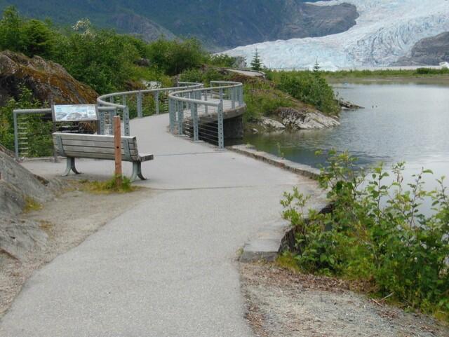A cement path winds approximately ten feet above an inlet of a calm lake, with a large glacier in the background