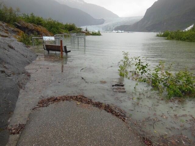 A cement path normally above a lake is completely submerged by grey floodwaters, with glacier visible in background.