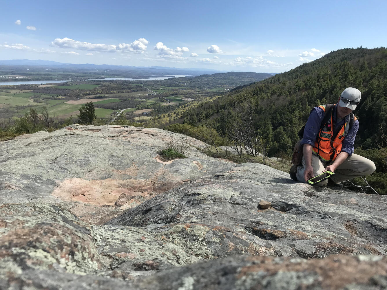 A researcher takes measurements from the top of Coot Hill. A lake and mountains can be seen in the background