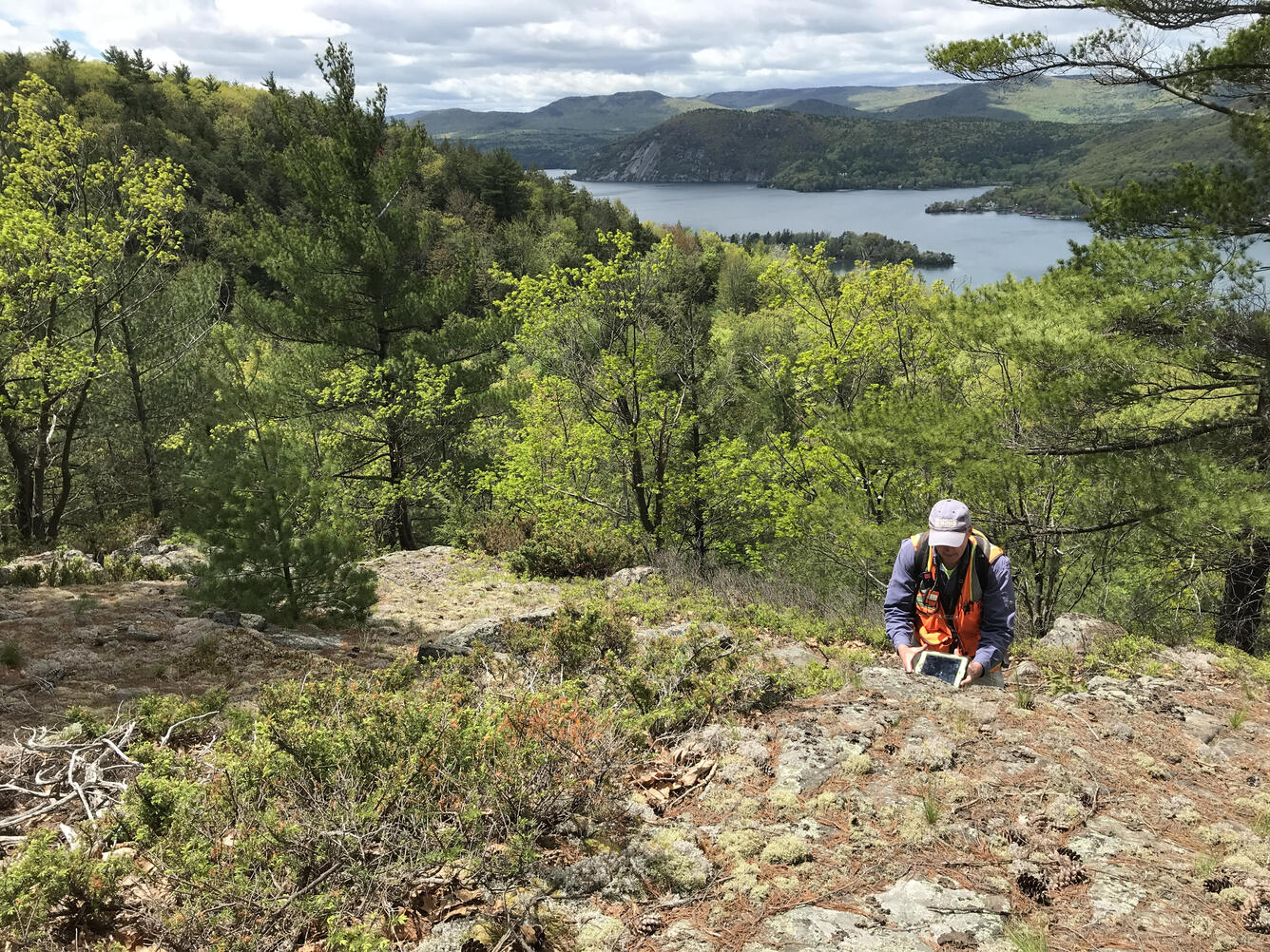 A researcher takes geologic measurements in the Adirondacks with Lake George and Rogers Rock in the distance