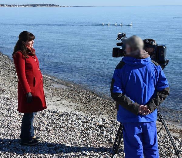 W woman in a red coat stands on the beach talking to a man in blue with a camera