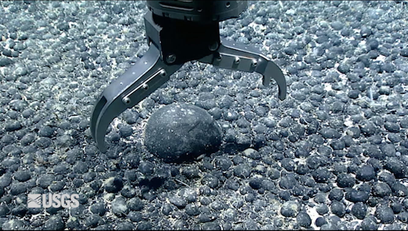 Close-up photograph showing a mechanical claw that is about to pick up a spherical rock off the seafloor.