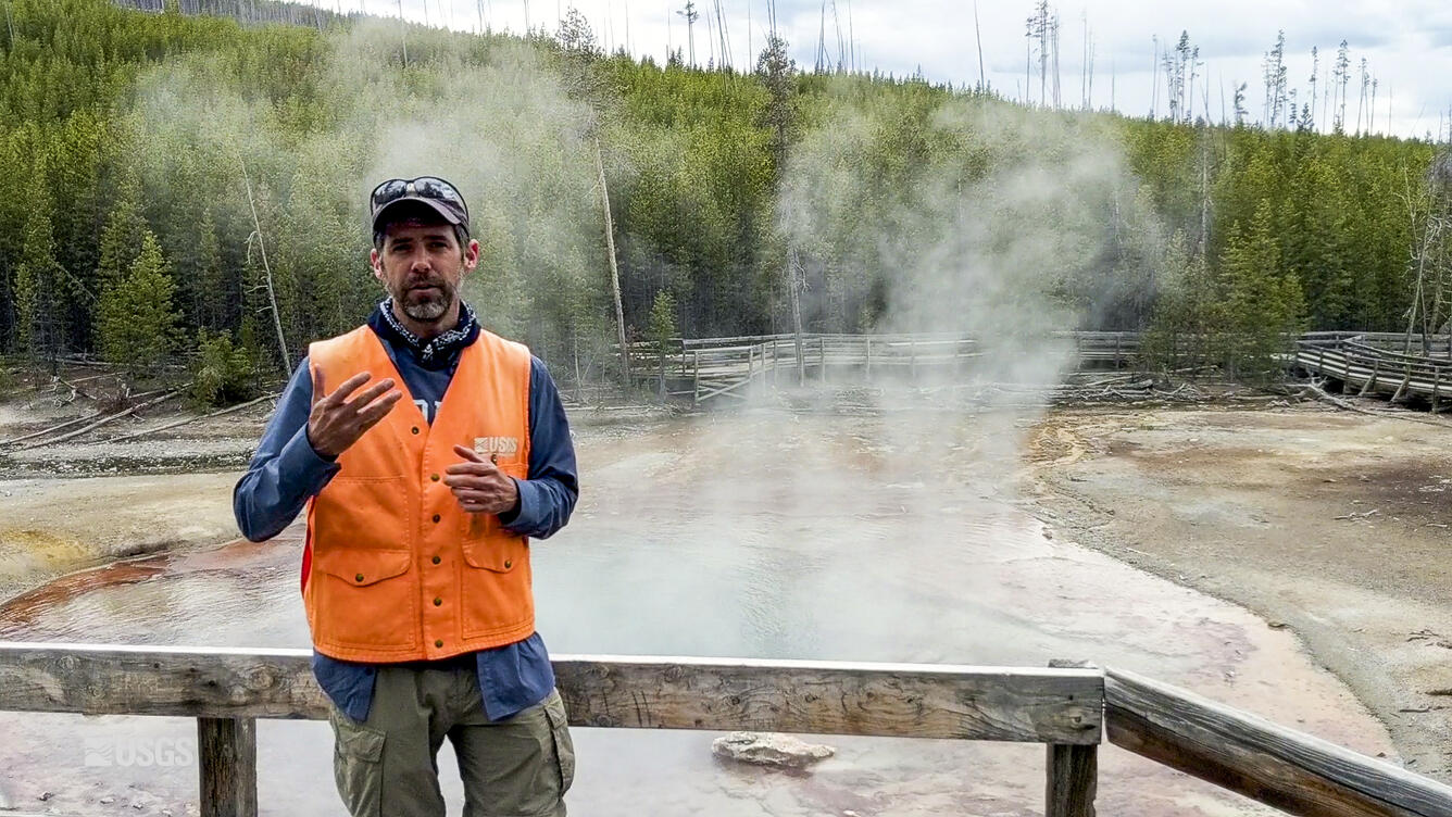 Man in orange safety jacket speaking while steam rises from a hot geyser behind him.