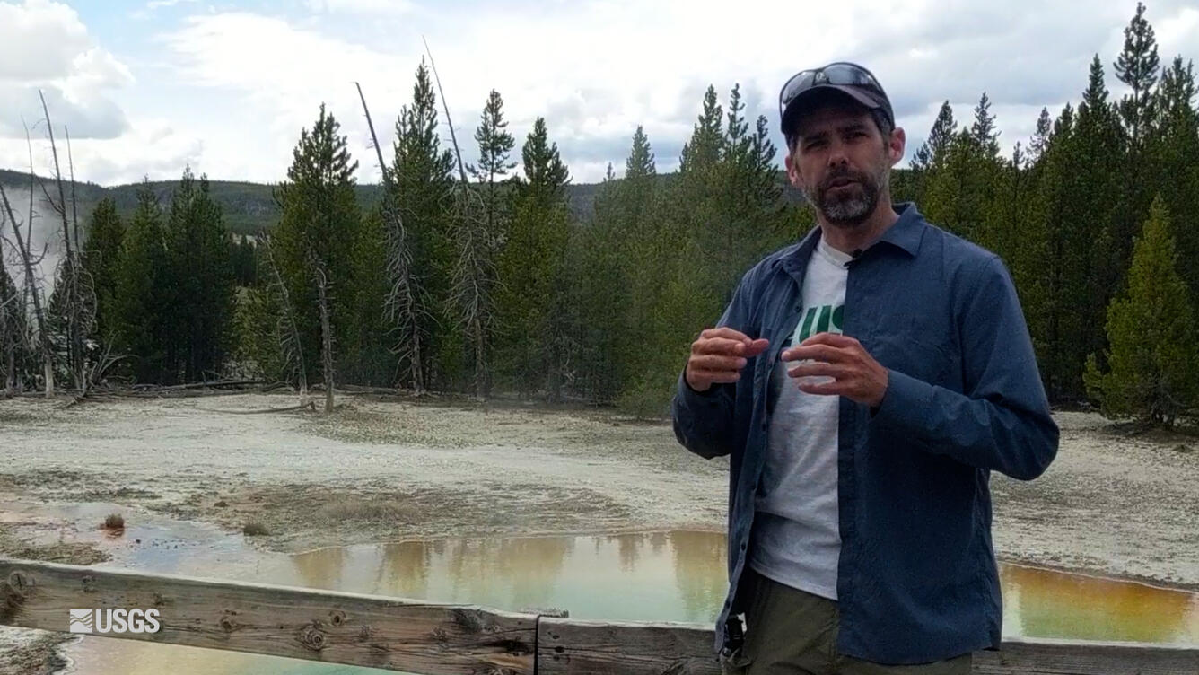 Man speaking in front of a small body of water, trees in the background.