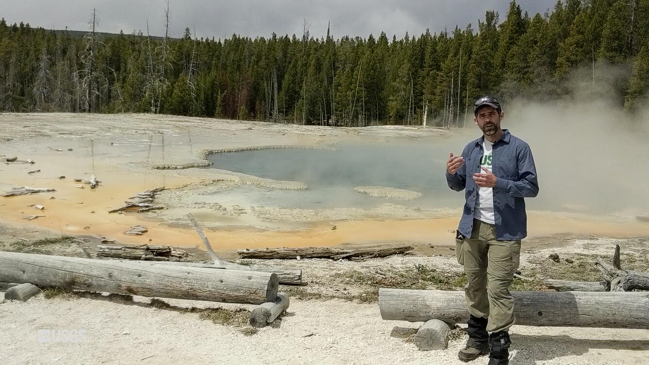 Man speaking in front of a small body of water, trees in the background.