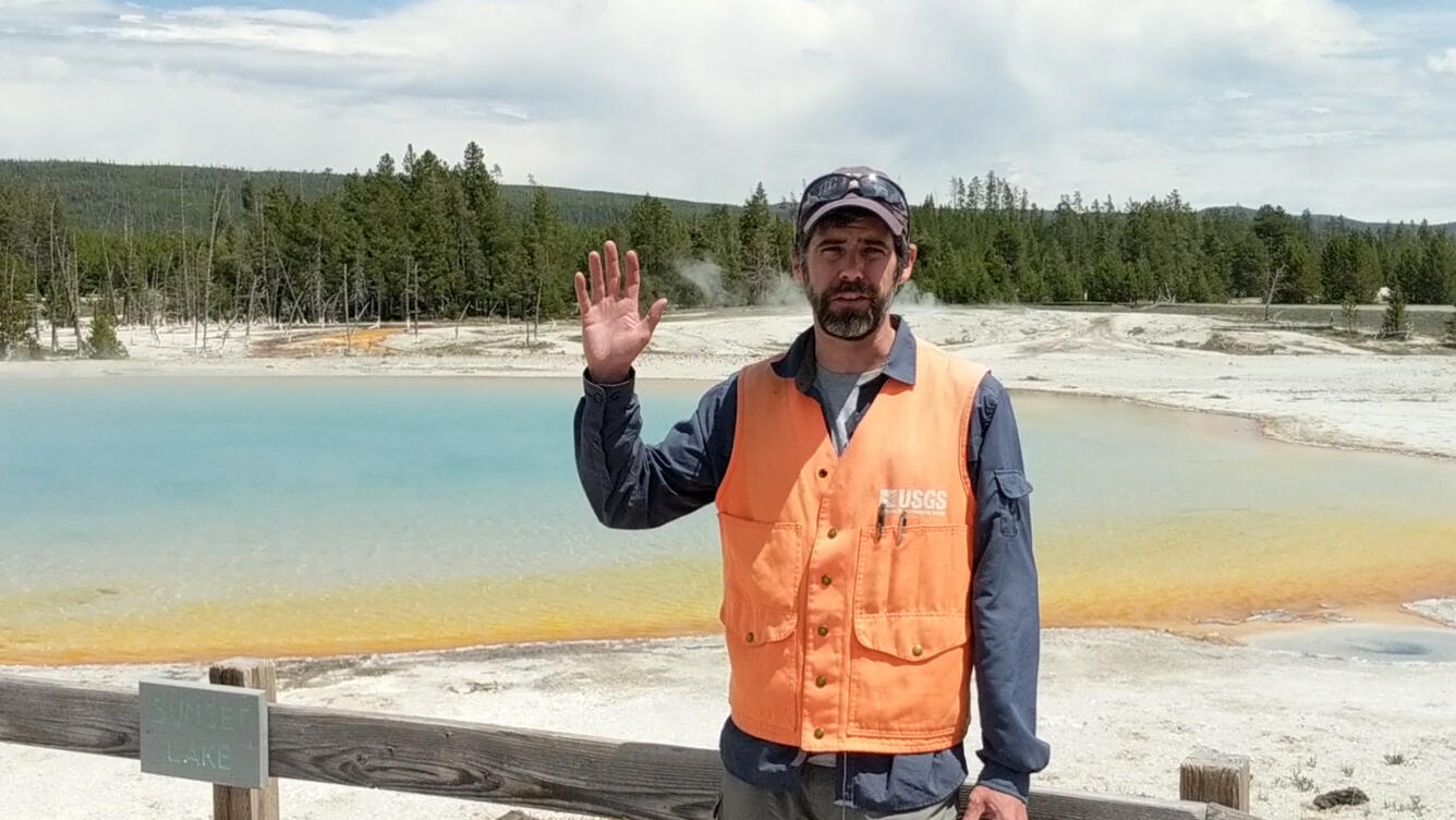 Man standing in front a water feature wearing an orange vest and waving hello with his hand.