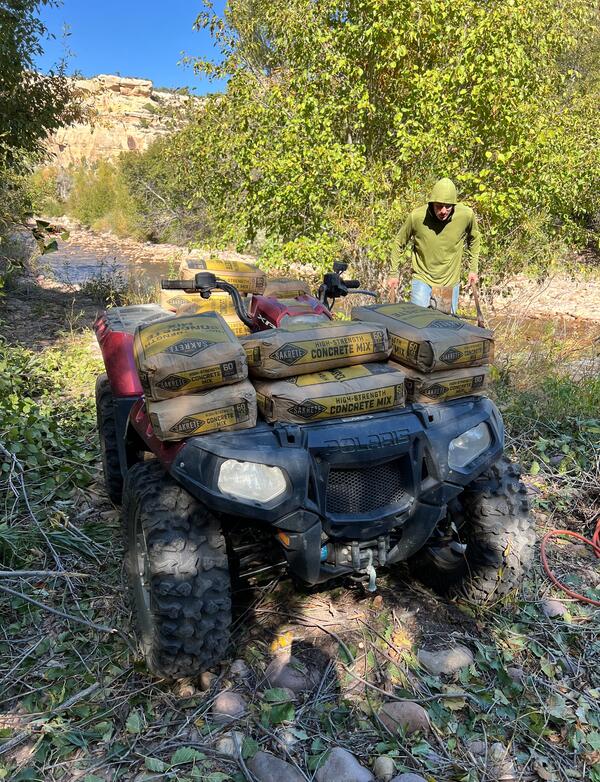 Four-runner loaded with bags of concrete is parked next to stream. Person on side behind the ATV.
