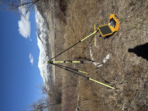 Surveying equipment is set up in a grassy field with snow-capped peaks in the distance.