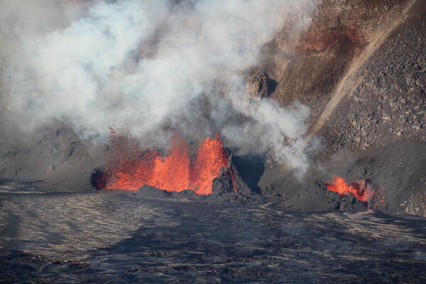 Orange lava fountains erupting inside of black lava spatter cone at the base of a crater wall