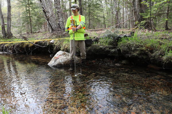 A man in neon shirt and life jacket stands in a creek holding a current meter.