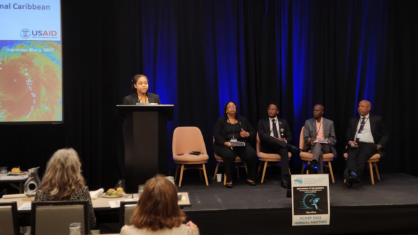 A woman speaks at a podium on stage in front of a crowd of people with a presentation about resilience in the Caribbean