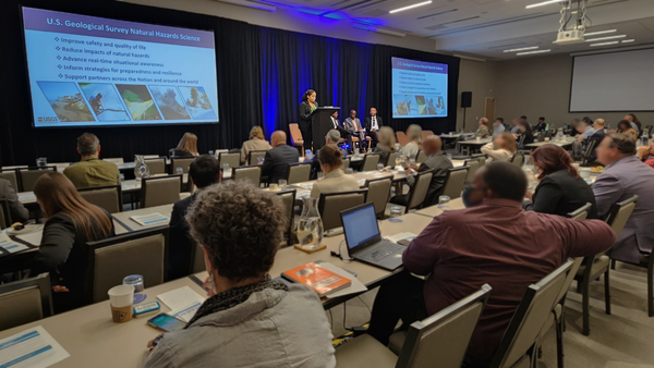 A woman speaks at a podium on stage in front of a crowd of people with a presentation about USGS natural hazards research