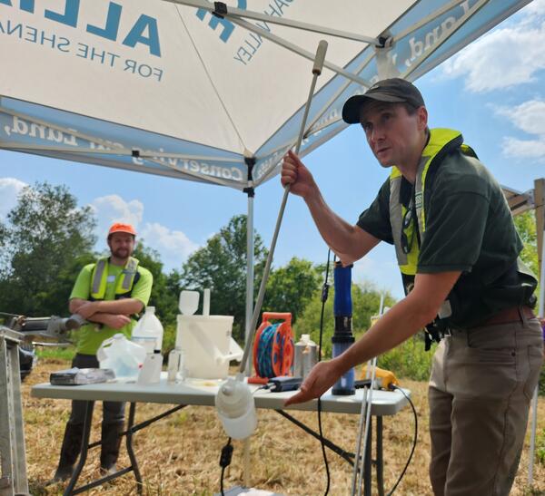 Two researchers in life vests stand at a table full of water quality sampling equipment. They demonstrate its use.