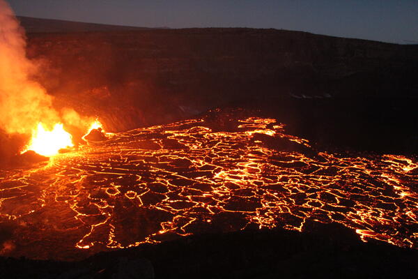 Low-light photo of yellow glowing lava fountains feeding red and black lava flows on crater floor