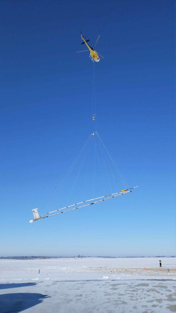 Helicopter towing large hoop over snow covered ground, technician for scale