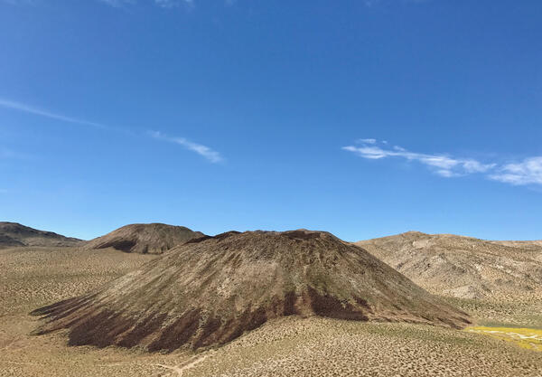 A photograph of several rocky volcanic hills shaped like upside-down flans. The hills and the landscape around them are dotted with sagebrush and dry grasses, revealing the underlying desert soil and rock. A bright blue sky overhead is streaked with clouds. USGS photo by Seth Burgess