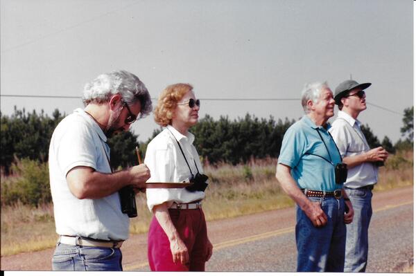 Three male and one female wearing binoculars for bird watching. One man is writing on a clipboard.