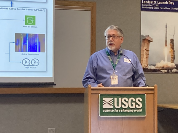 A man speaks at a wood podium with a large screen to his right and a poster behind him.
