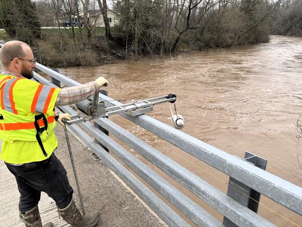 Hydrologic technician collects water sample from flooded river while standing on a bridge