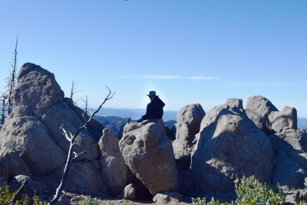 A scientist in a broad-brimmed hat sits on a large gray boulder of volcanic rock, surrounded by similar boulders. They are holding a rock hammer against the rock in preparation to break off a sample. In the distance, several hazy mountain ridges can be seen. 