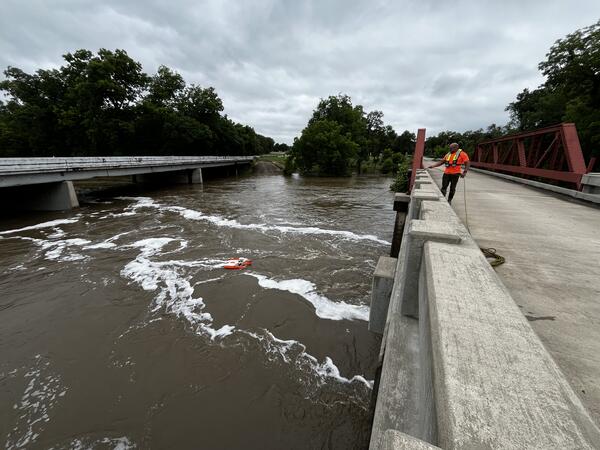 A water specialist stands on a bridge holding a line that controls orange floating water monitoring equipment in the fast-mov
