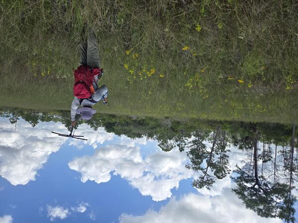 a person with red backpack and purple hat holds up a tracking device in a field with yellow flowers, trees in background