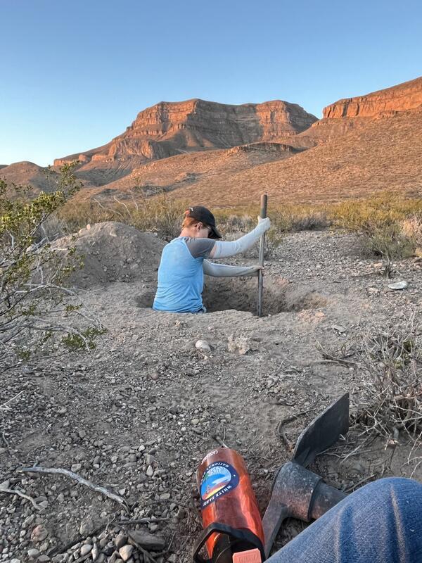 Person standing in a small diameter hole above their waist while digging with a shovel. Mountains in background.