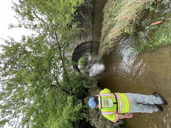 A USGS technician in a bright green safety jacket takes a photo of a flooded river