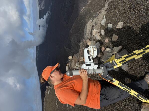 Color photograph of field engineer servicing a livestream camera with volcanic vents degassing in the background