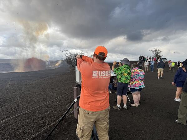 Color photograph of scientist setting up equipment with visitors viewing a volcanic eruption nearby