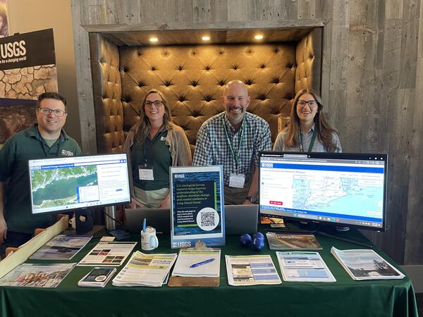 Group of USGS scientists smiling for photo behind booth with handouts and two computer monitors displaying maps