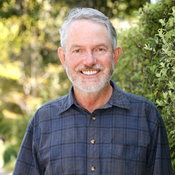 A man wearing a blue collared shirt standing outside with trees in the background