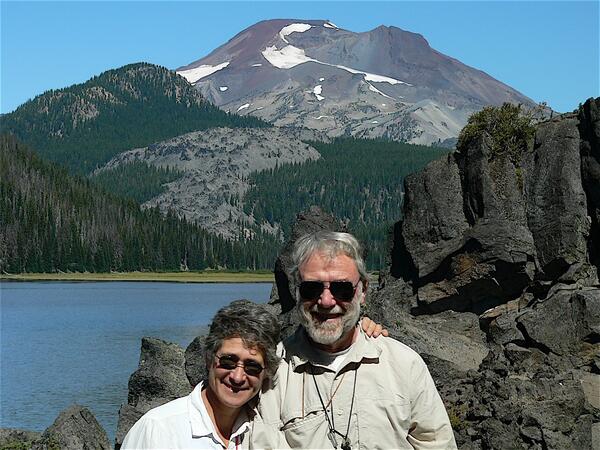 An older man and woman standing in front of a rock outcrop with a lake and forested volcano in the background. Both are wearing khaki shirts, sunglasses, and hand lenses. 