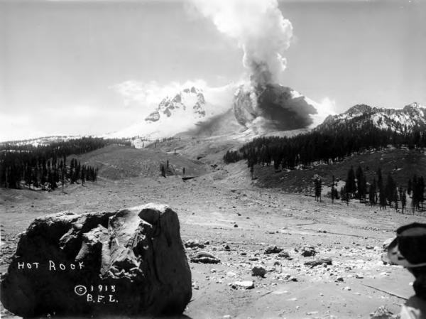 A black and white photograph looks across a swath cut through a pine forest by a debris and mud flow that originates at a steaming volcanic peak in the distance. A large crater is visible on the side of the volcano, which is emitting the steam. The rest of the volcano is heavily blanketed in snow.