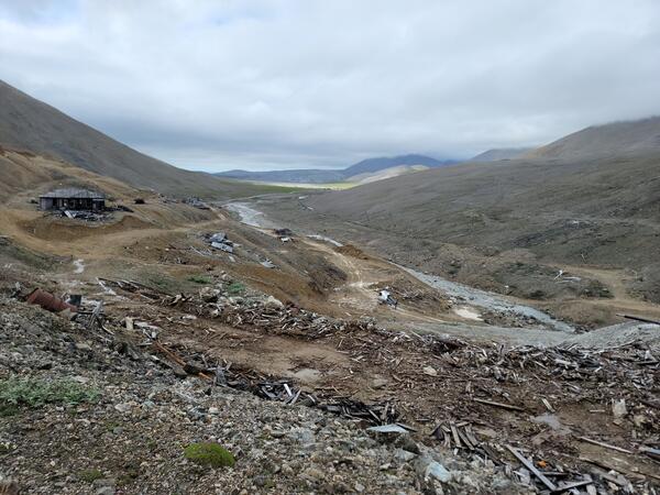 Old buildings, rusted barrel, rocks, and scattered wood from previous buildings in mountain valley. Helicopter near stream.