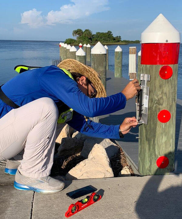 Image shows a USGS scientist in a PFD installing a storm-tide sensor on a pier