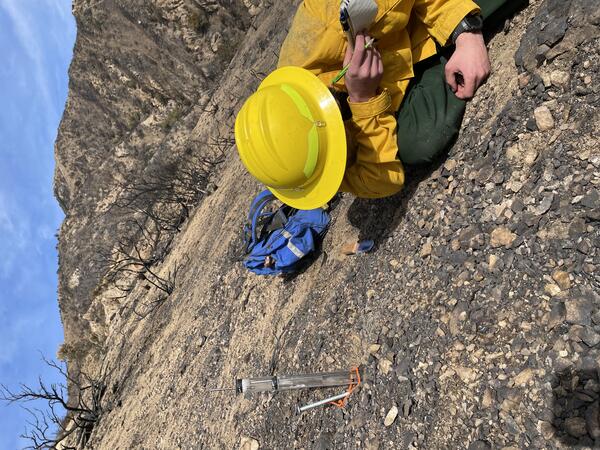 person holding notebook and wearing hardhat lies on ground watching graduated cylinder and recording measurements