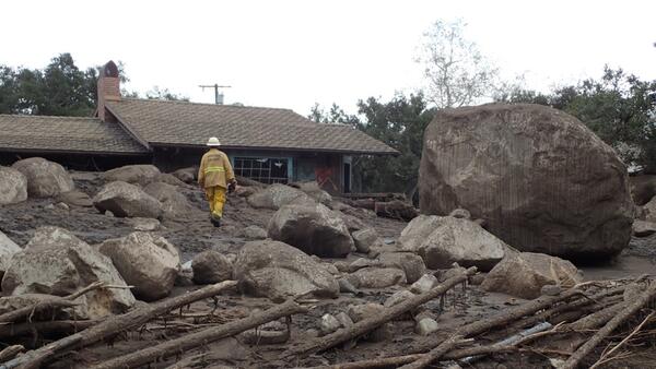 Large boulders and person walking in front of damaged house