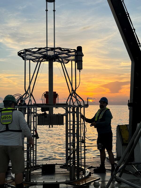 two people working with a mega multicorer on the deck of a ship at sunset