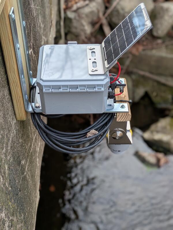 Close up view of gray box with solar panel affixed to bridge above stream