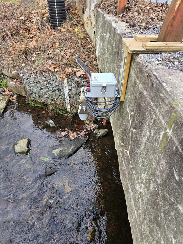 Gray box with attached solar panel attached to bridge with stream below
