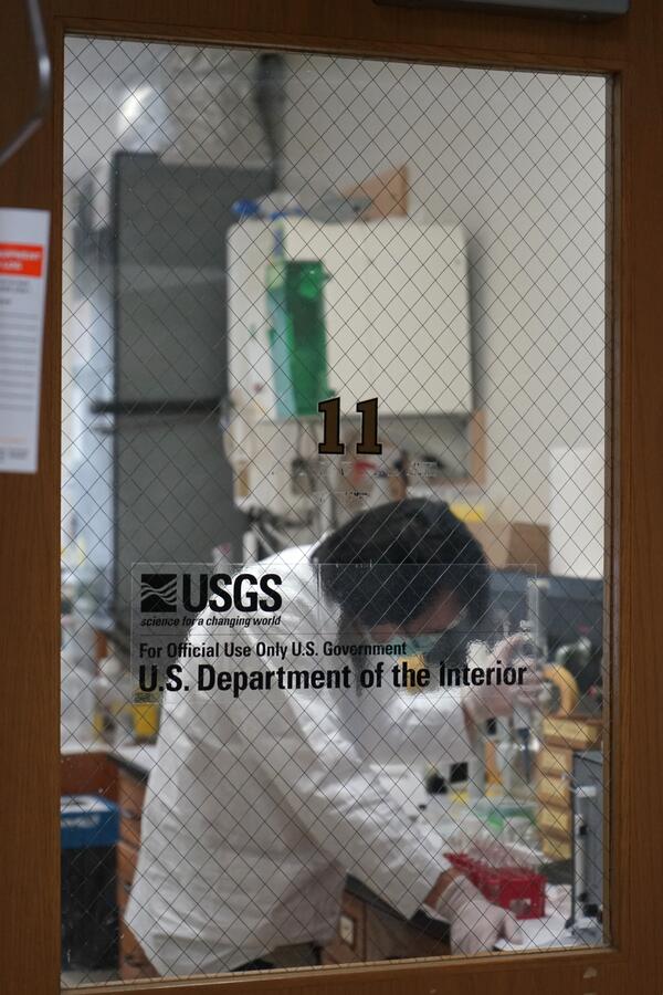 View of the lab from the outside, looking through glass window at scientist prepping samples