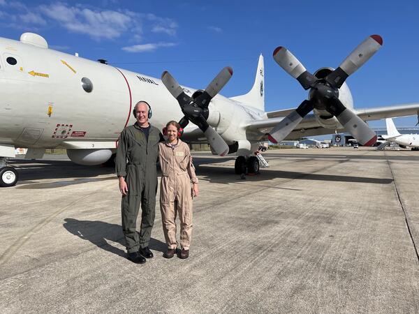 Two people in flight suits standing together smiling next to an aircraft on a runway